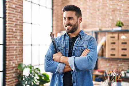 Young hispanic man smiling confident standing with arms crossed gesture at art studio