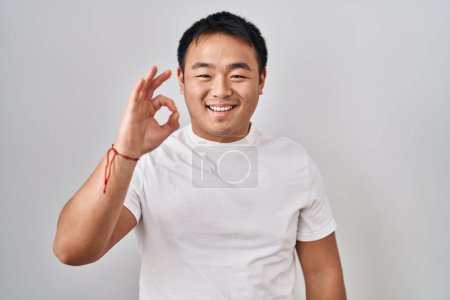 Téléchargez les photos : Jeune homme chinois debout sur fond blanc souriant positif faire ok signe avec la main et les doigts. expression réussie. - en image libre de droit