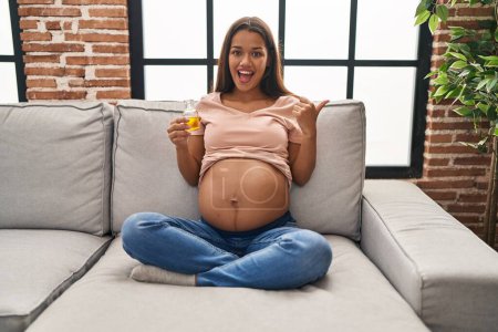 Foto de Mujer embarazada joven sosteniendo aceite hidratante para hidratar el vientre apuntando el pulgar hacia el lado sonriendo feliz con la boca abierta - Imagen libre de derechos