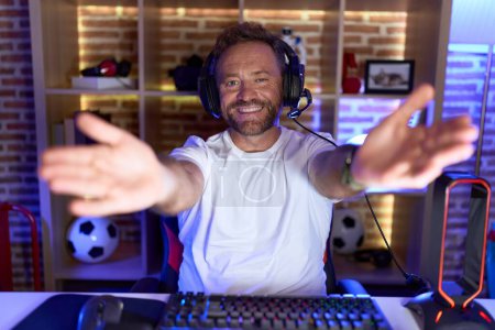 Photo for Middle age man with beard playing video games wearing headphones looking at the camera smiling with open arms for hug. cheerful expression embracing happiness. - Royalty Free Image