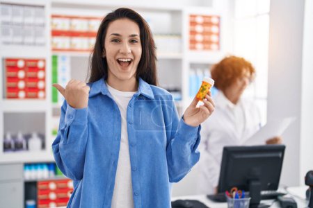 Foto de Joven morena sosteniendo pastillas en la farmacia apuntando con el pulgar hacia el lado sonriendo feliz con la boca abierta - Imagen libre de derechos