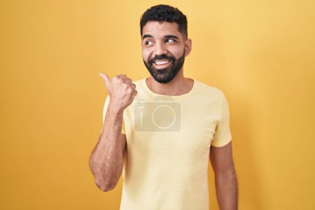 Photo for Hispanic man with beard standing over yellow background smiling with happy face looking and pointing to the side with thumb up. - Royalty Free Image