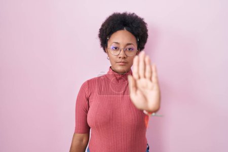Téléchargez les photos : Belle femme africaine avec les cheveux bouclés debout sur fond rose faisant arrêter de chanter avec la paume de la main. expression d'avertissement avec geste négatif et sérieux sur le visage. - en image libre de droit