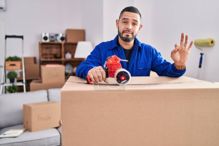 Photo for Hispanic man working on moving holding packing tape doing ok sign with fingers, smiling friendly gesturing excellent symbol - Royalty Free Image
