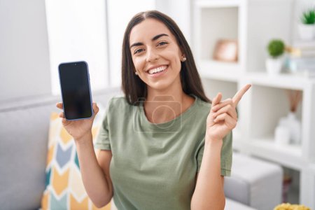 Foto de Young brunette woman holding smartphone showing blank screen smiling happy pointing with hand and finger to the side - Imagen libre de derechos