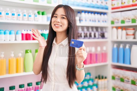 Foto de Joven mujer china de compras en farmacia sosteniendo tarjeta de crédito celebrando la victoria con sonrisa feliz y la expresión del ganador con las manos levantadas - Imagen libre de derechos