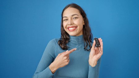 Téléchargez les photos : Jeune belle femme hispanique souriant confiant pointant vers la clé de la nouvelle voiture sur fond bleu isolé - en image libre de droit