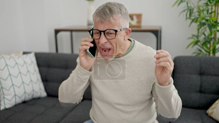 Foto de Hombre de pelo gris de mediana edad sentado en el sofá con expresión furiosa hablando en el teléfono inteligente en casa - Imagen libre de derechos