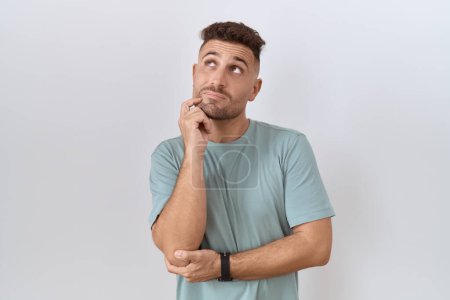 Foto de Hispanic man with beard standing over white background with hand on chin thinking about question, pensive expression. smiling with thoughtful face. doubt concept. - Imagen libre de derechos