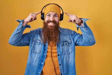Foto de Hombre caucásico con barba larga escuchando música usando auriculares sonriendo apuntando a la cabeza con ambas manos dedo, gran idea o pensamiento, buena memoria - Imagen libre de derechos