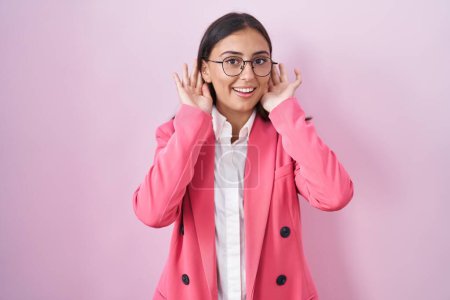 Foto de Mujer hispana joven vestida con ropa de negocios y gafas tratando de escuchar ambos gestos de manos en la oreja, curiosa de chismes. problema auditivo, sordo - Imagen libre de derechos