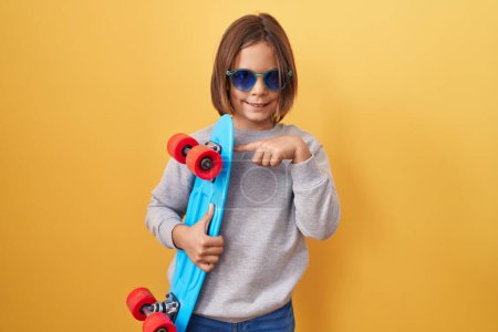 Foto de Pequeño niño hispano con gafas de sol sosteniendo patinaje sonriendo feliz señalando con la mano y el dedo - Imagen libre de derechos