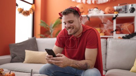 Foto de Joven hombre caucásico con traje de diablo sentado en el sofá usando el teléfono inteligente en casa - Imagen libre de derechos