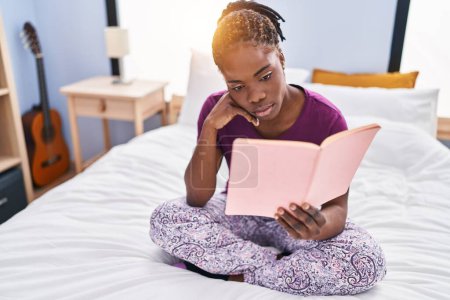 Foto de African american woman reading book sitting on bed at bedroom - Imagen libre de derechos