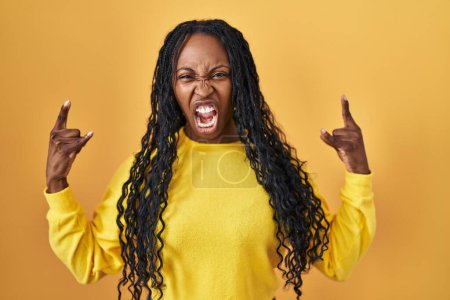 Photo for African woman standing over yellow background shouting with crazy expression doing rock symbol with hands up. music star. heavy concept. - Royalty Free Image