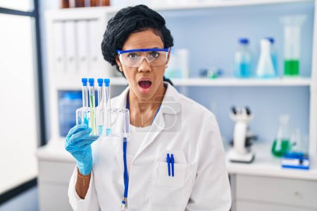 Photo for Middle age hispanic woman working at scientist laboratory scared and amazed with open mouth for surprise, disbelief face - Royalty Free Image
