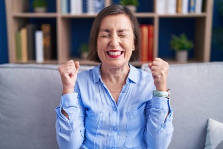 Photo for Middle age hispanic woman sitting on the sofa at home excited for success with arms raised and eyes closed celebrating victory smiling. winner concept. - Royalty Free Image