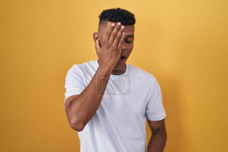 Photo for Young hispanic man standing over yellow background yawning tired covering half face, eye and mouth with hand. face hurts in pain. - Royalty Free Image