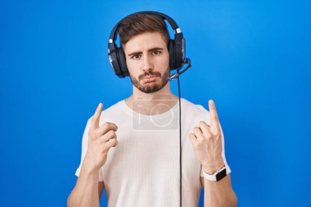 Foto de Hombre hispano con barba escuchando música con auriculares apuntando hacia arriba, mirando triste y molesto, indicando dirección con los dedos, infeliz y deprimido. - Imagen libre de derechos