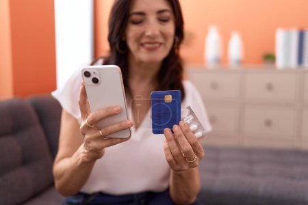 Photo for Middle age hispanic woman using smartphone and credit card sitting on sofa at home - Royalty Free Image