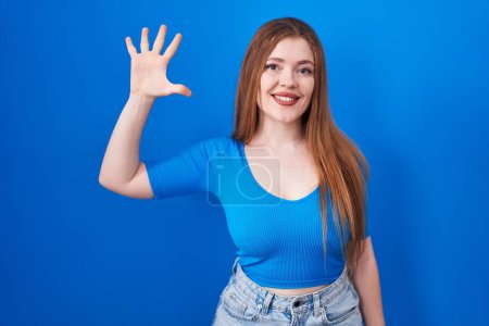 Téléchargez les photos : Femme rousse debout sur fond bleu montrant et pointant vers le haut avec les doigts numéro cinq tout en souriant confiant et heureux. - en image libre de droit