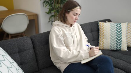 Photo for Young blonde woman writing on notebook sitting on sofa at home - Royalty Free Image