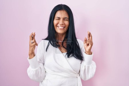 Téléchargez les photos : Femme hispanique mature debout sur fond rose doigt gestuel croisé souriant avec espoir et les yeux fermés. chance et concept superstitieux. - en image libre de droit
