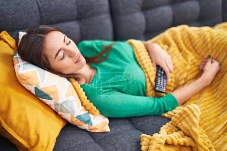Photo for Young beautiful hispanic woman holding tv remote control lying on sofa sleeping at home - Royalty Free Image