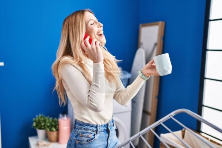 Téléchargez les photos : Young blonde woman talking on smartphone drinking coffee waiting for washing machine at laundry room - en image libre de droit