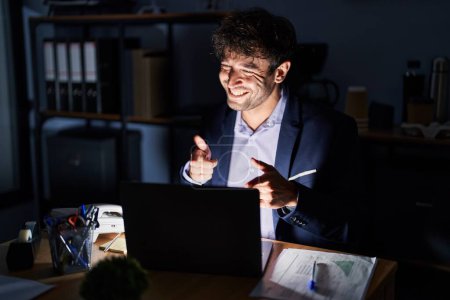 Photo for Hispanic young man working at the office at night pointing fingers to camera with happy and funny face. good energy and vibes. - Royalty Free Image