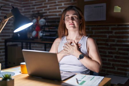 Téléchargez les photos : Femme brune travaillant au bureau la nuit souriant avec les mains sur la poitrine avec les yeux fermés et geste reconnaissant sur le visage. concept de santé. - en image libre de droit