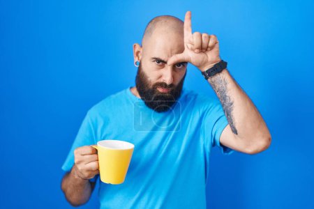Téléchargez les photos : Jeune homme hispanique avec barbe et tatouages boire une tasse de café se moquer des gens avec les doigts sur le front faire geste de perdant moqueur et insultant. - en image libre de droit