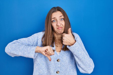 Foto de Young woman standing over blue background doing thumbs up and down, disagreement and agreement expression. crazy conflict - Imagen libre de derechos