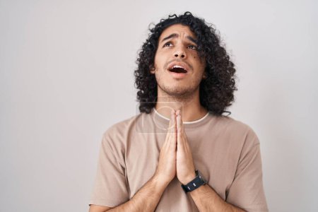 Téléchargez les photos : Homme hispanique avec les cheveux bouclés debout sur fond blanc mendier et prier avec les mains ainsi que l'expression de l'espoir sur le visage très émotionnel et inquiet. mendier. - en image libre de droit