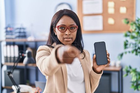 Téléchargez les photos : Jeune femme africaine tenant smartphone montrant écran vide pointant du doigt à la caméra et à vous, geste confiant regardant sérieux - en image libre de droit