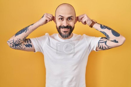 Téléchargez les photos : Jeune homme hispanique avec des tatouages debout sur fond jaune souriant tirant les oreilles avec les doigts, geste drôle. problème d'audition - en image libre de droit