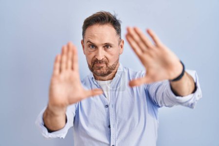 Photo for Middle age caucasian man standing over blue background doing frame using hands palms and fingers, camera perspective - Royalty Free Image