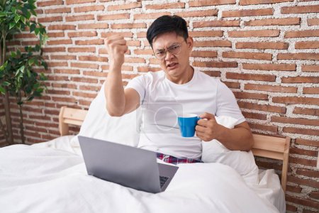Foto de Joven asiático bebiendo café sentado en la cama molesto y frustrado gritando con ira, gritando loco de ira y la mano levantada - Imagen libre de derechos