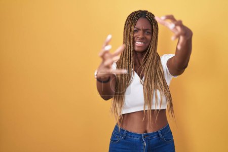 Téléchargez les photos : Femme afro-américaine avec les cheveux tressés debout sur fond jaune criant frustré par la rage, les mains essayant d'étrangler, hurlant fou - en image libre de droit