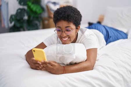 Foto de African american woman using smartphone lying on bed at bedroom - Imagen libre de derechos