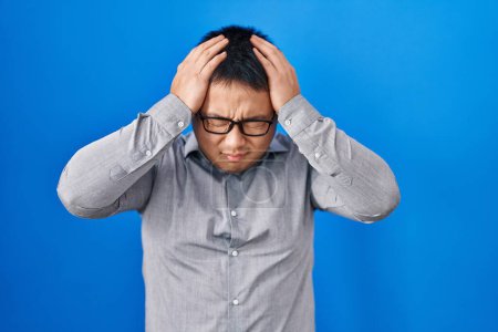 Téléchargez les photos : Jeune homme chinois debout sur fond bleu souffrant de maux de tête désespérés et stressés parce que la douleur et la migraine. mains sur la tête. - en image libre de droit