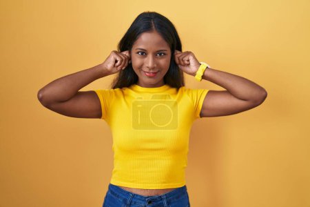Foto de Mujer india joven de pie sobre fondo amarillo sonriendo tirando de las orejas con los dedos, gesto divertido. problema de audición - Imagen libre de derechos