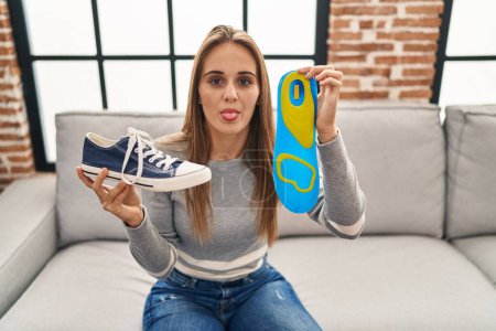Foto de Mujer joven sosteniendo plantilla de zapato con la lengua hacia fuera feliz con expresión divertida. - Imagen libre de derechos