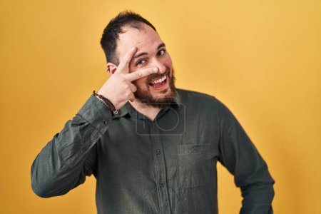 Téléchargez les photos : Plus taille homme hispanique avec barbe debout sur fond jaune faisant symbole de paix avec les doigts sur le visage, souriant joyeux montrant la victoire - en image libre de droit