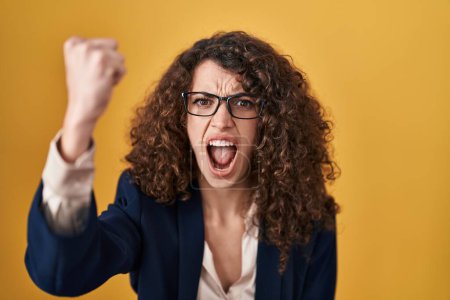 Photo for Hispanic woman with curly hair standing over yellow background angry and mad raising fist frustrated and furious while shouting with anger. rage and aggressive concept. - Royalty Free Image