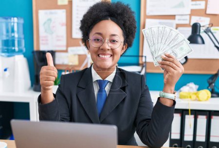 Téléchargez les photos : Belle femme africaine avec les cheveux bouclés tenant de l'argent au bureau souriant heureux et positif, pouce levé faire excellent et signe d'approbation - en image libre de droit
