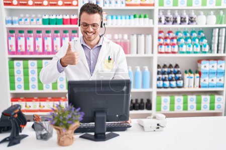 Téléchargez les photos : Jeune homme hispanique travaillant à la pharmacie pharmacie portant casque souriant heureux et positif, pouce levé faire excellent et signe d'approbation - en image libre de droit