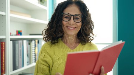 Photo for Middle age hispanic woman teacher smiling confident reading book at library university - Royalty Free Image