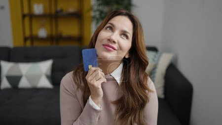 Foto de Joven mujer hispana hermosa sosteniendo tarjeta de crédito sentada en el sofá en casa - Imagen libre de derechos