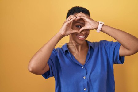 Photo for African american woman standing over yellow background doing heart shape with hand and fingers smiling looking through sign - Royalty Free Image